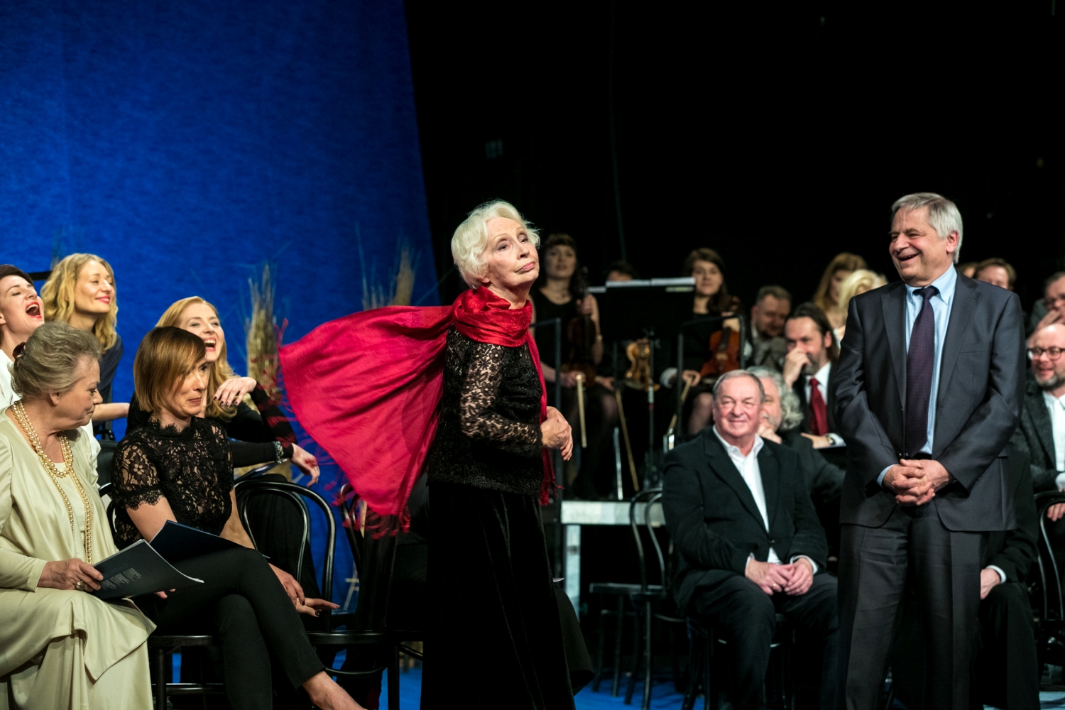 Scena zbiorowa, starsza kobieta ubrana na czarno, z rozwianym czerwonym szalem, stoi na scenie.