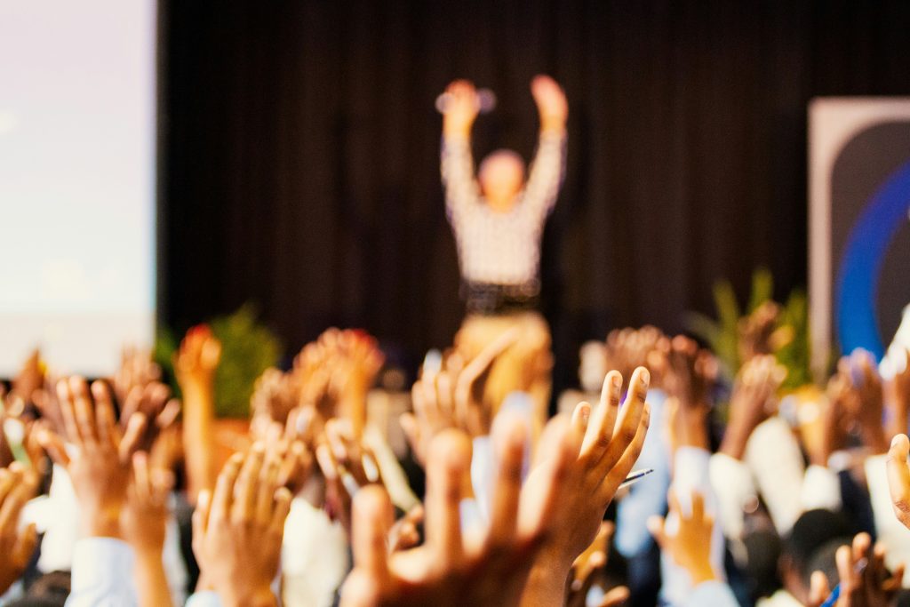 Zróżnicowana grupa osób stojących tyłem podnoszących ręce do góry, widok uniesionych dłoni. W tle zarys sylwetki jednej osoby stojącej przodem do grupy z wyciągniętymi do góry rękami. 