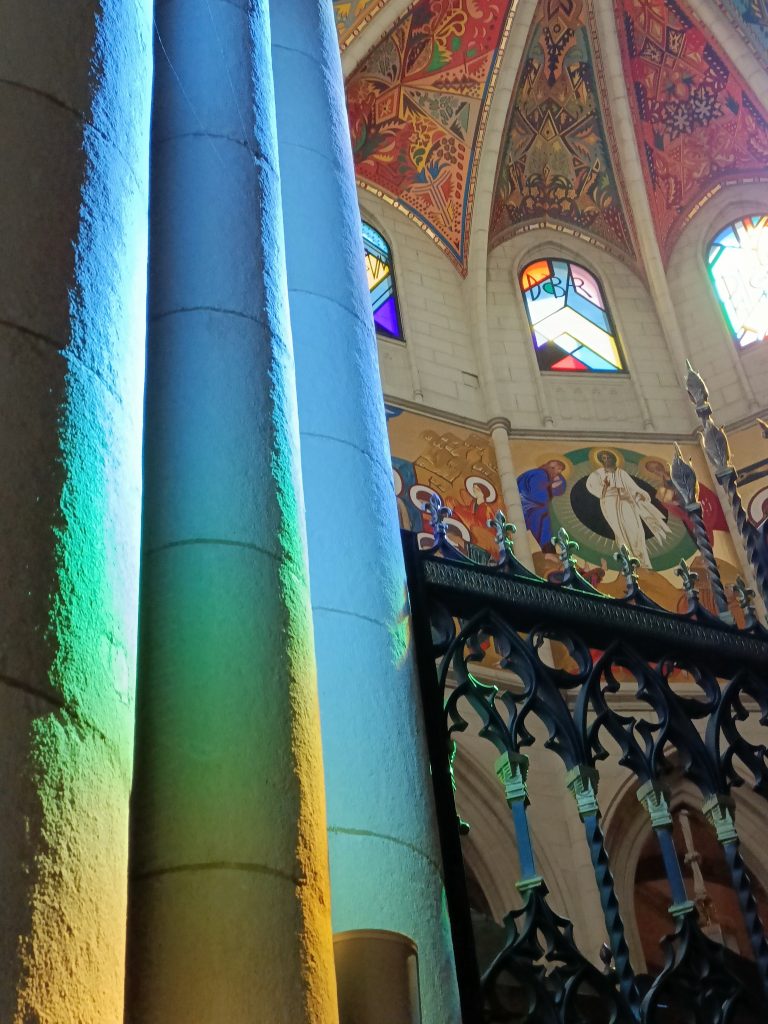 Żywe kolorowe wnętrze kościoła, kolorowe polichromie na ścianach i sklepieniu, w oknach kolorowe witraże. Na pierwszym planie kolumna, na której odbija się tęczowe światło witraży.