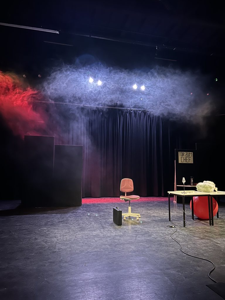 Czarna sala, na środku sceny krzesło na kółkach, obok czarna aktówka. Po prawej stronie stolik, duża czerwona dmuchana piłka. Nad sceną unoszą się czerwone i białe dymy.