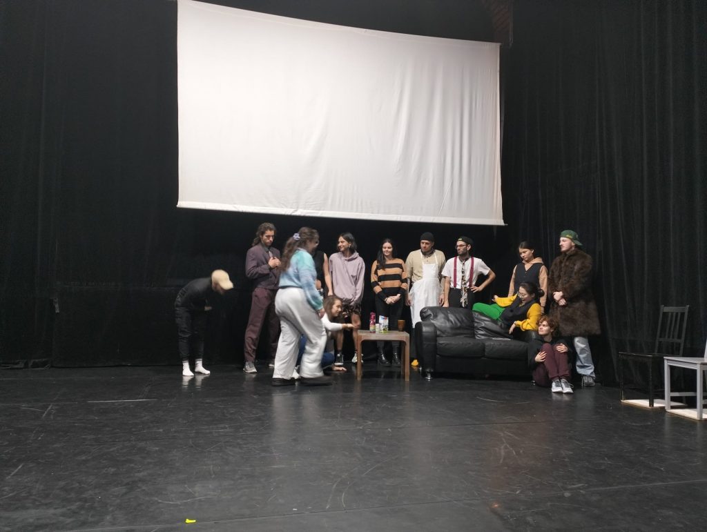 Zróżnicowana grupa ludzi zgromadzona na scenie, po prawej stronie czarna skórzana kanapa, drewniany stolik. Nad nimi wisi biały ekran do projektora.